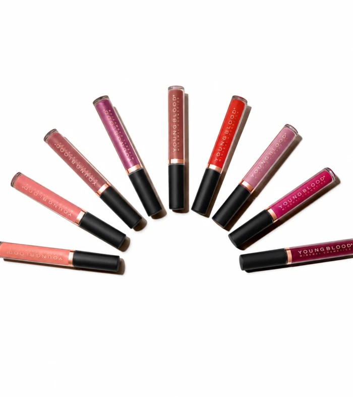 Assorted Youngblood lip gloss tubes arranged in a curved arc on a white background, showing various pinks, reds, and nudes.