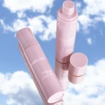 Pink sunscreen tubes floating against a bright blue sky with white clouds, showcasing the product shape and cap design.