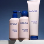 Three pink Darling body care bottles with blue pump and cap against a blue gradient background on a shelf, branding visible (Darling).