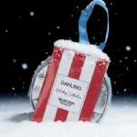 Red and white striped gift bag with snow on top, blue handle, leaning against silver headphones in a snowy scene; reads 'Darling Holiday Collection'.
