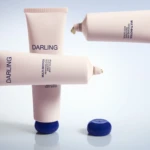 Three pale pink Darling body scrub tubes arranged diagonally on a reflective surface, with blue caps off to the side; text on the tubes reads 'DARLING' and 'MULTI-BIOTICS Renewing and Smoothing Body Scrub'