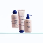 Three pastel pink Darling skincare bottles and pump tube arranged on a glass shelf with a blue cap and string decor, against a white background