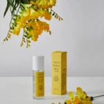 Yellow freesia flowers hang over a white surface beside yellow perfume packaging and a spray bottle labeled Lemon Freesia Musk.