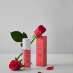 Pink perfume bottle labeled Lychee Rose Cedar beside its pink box, with a red rose and petals on a white surface.
