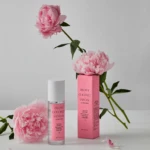 Pink peonies with a pink perfume bottle and matching box arranged on a white surface against a light gray background.