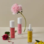 Flat lay of pink peony with three fragrance bottles (Lychee Rose Cedar, Peony Coconut Sandal, Lemon Freesia Musk) on a pale surface, with scattered petals and a red rose at left.