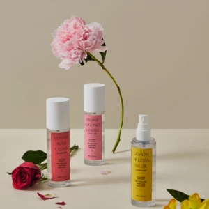 Flat lay of pink peony with three fragrance bottles (Lychee Rose Cedar, Peony Coconut Sandal, Lemon Freesia Musk) on a pale surface, with scattered petals and a red rose at left.