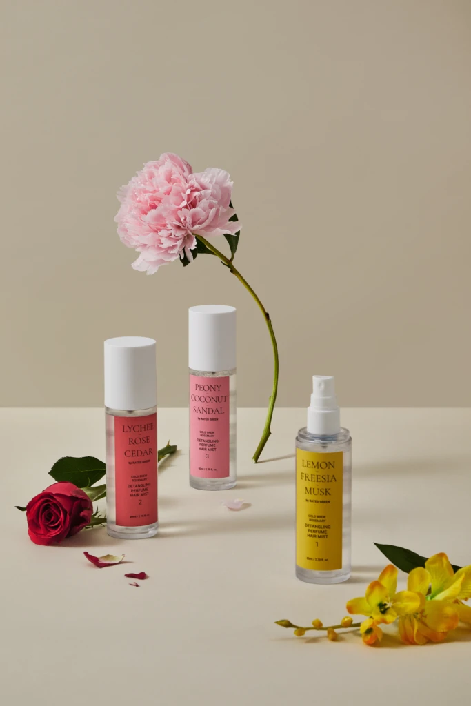 Flat lay of pink peony with three fragrance bottles (Lychee Rose Cedar, Peony Coconut Sandal, Lemon Freesia Musk) on a pale surface, with scattered petals and a red rose at left.