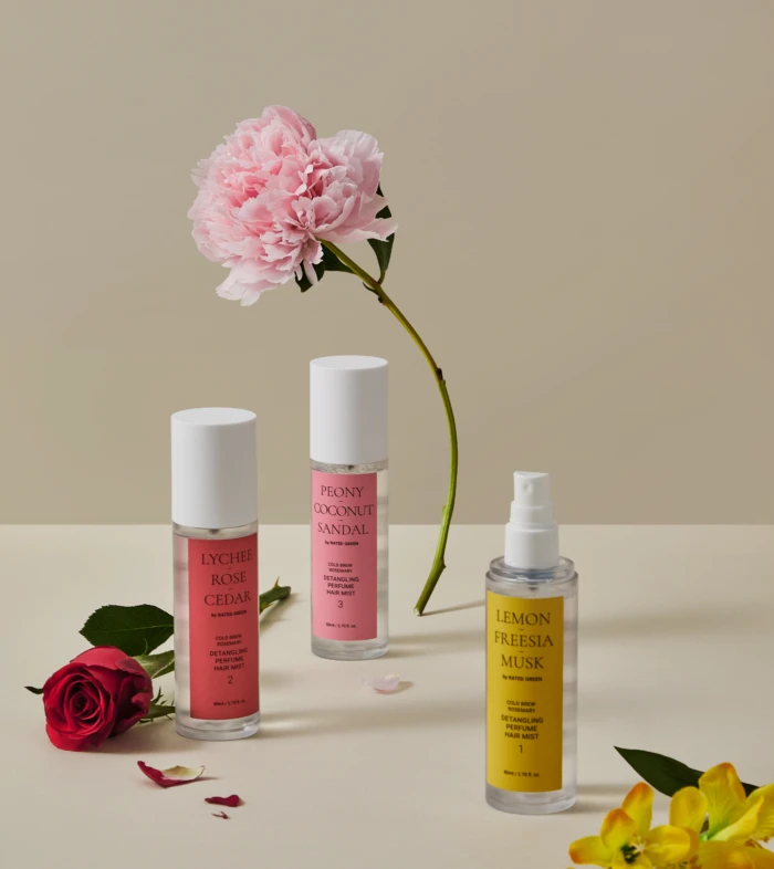 Flat lay of pink peony with three fragrance bottles (Lychee Rose Cedar, Peony Coconut Sandal, Lemon Freesia Musk) on a pale surface, with scattered petals and a red rose at left.