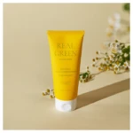 Yellow Real Green Natural Kids Conditioner tube standing upright on a beige surface with small white flowers in the background.