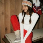Woman in a white long-sleeve top and red ski pants sits on a bench, wearing a knit headband that reads 'Ski Club' with braided hair and a smile.