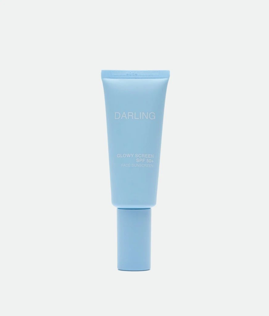 Blue sunscreen tube labeled 'DARLING' with SPF 50+ standing upright against a pale background, showing 'GLOWY SCREEN' text.