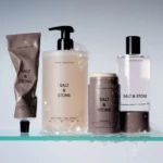 Assorted Salt & Stone bathroom products on a glass shelf: body wash, lotion, deodorant, perfume-like bottle, and a tube, all with minimalistic black lettering against pale containers and a soft blue-gray background
