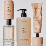 Beige Salt & Stone skincare set: pump bottle, deodorant, lotion, and tube with water droplets on a reflective surface.