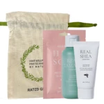 Beige fabric tote with green strap containing a hair care gift set: pink pouch, teal bottle, and white Real Shea tube.