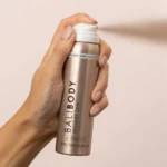 Hand gripping a metallic spray can labeled BaliBody Instant Tan, spraying a fine mist to the right.
