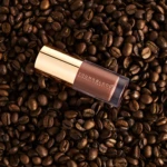Lip gloss or liquid makeup tube resting on dark roasted coffee beans, branding reads Youngblood Mineral Cosmetics.
