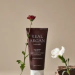Brown Real Argan hair mask tube with a red flower leaning beside it on a beige background, minimalist style.