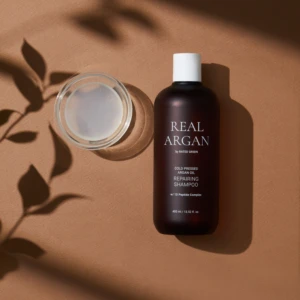 Brown bottle labeled Real Argan shampoo with a white cap, placed next to a glass jar of cream on a warm brown surface, with leaf shadows nearby.