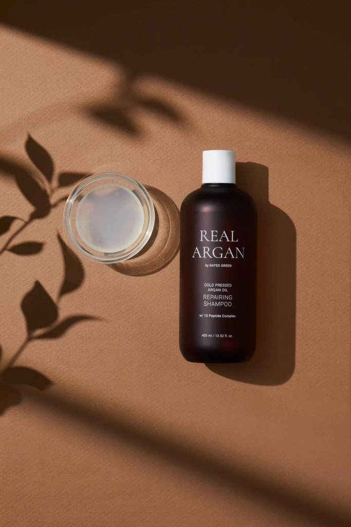 Brown bottle labeled Real Argan shampoo with a white cap, placed next to a glass jar of cream on a warm brown surface, with leaf shadows nearby.
