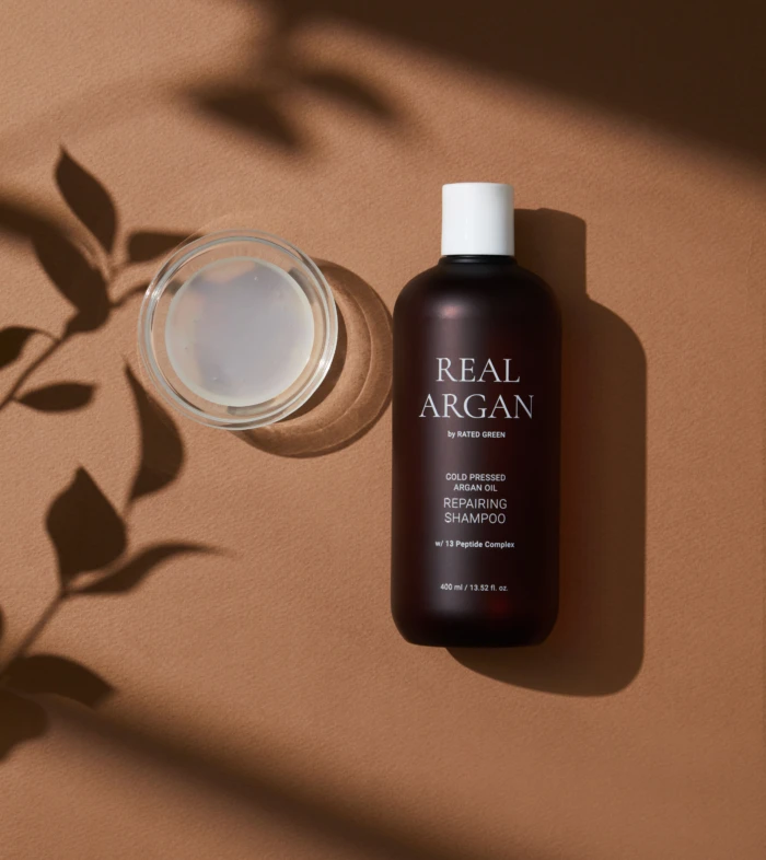 Brown bottle labeled Real Argan shampoo with a white cap, placed next to a glass jar of cream on a warm brown surface, with leaf shadows nearby.