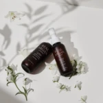 Two brown bottles with white pumps labeled Real Argan hair oil, resting on a white surface among white flowers and soft shadows.