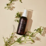 Brown bottle of Real ArgAN Smoothing Hair Serum with a white pump, laid on a pale beige background among white flowers and green stems.