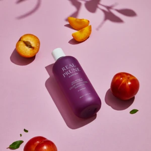 Purple Real Prune shampoo bottle on a pink surface with apricot slices and whole peaches nearby.