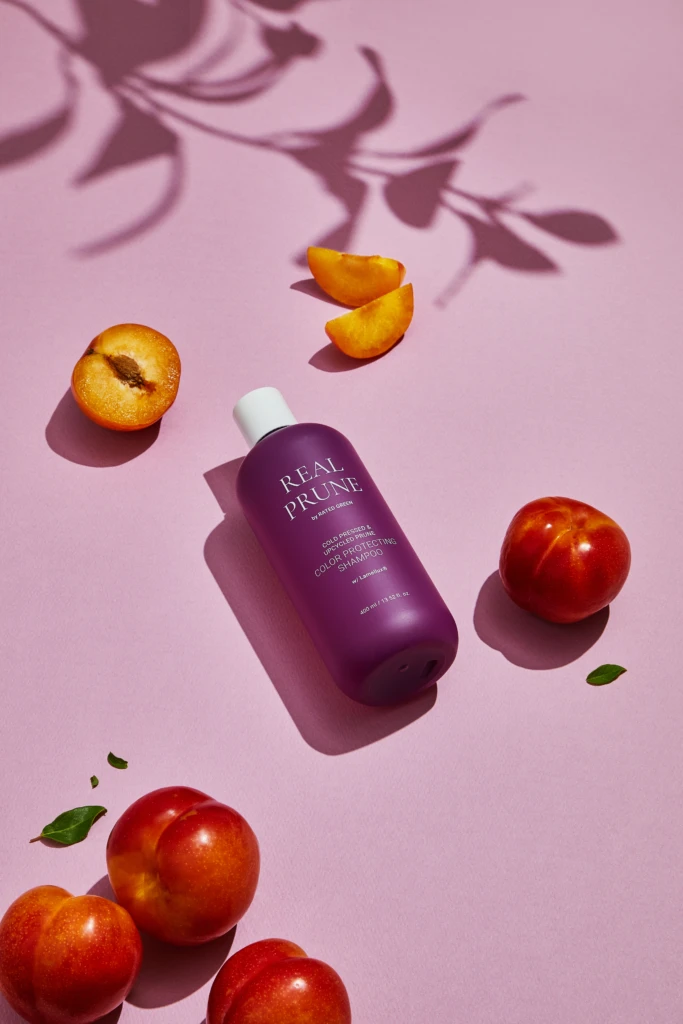 Purple Real Prune shampoo bottle on a pink surface with apricot slices and whole peaches nearby.