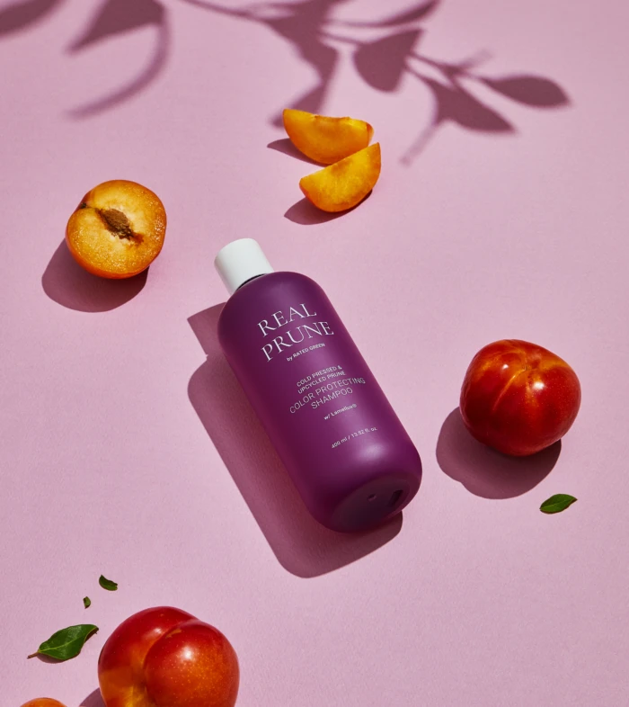 Purple Real Prune shampoo bottle on a pink surface with apricot slices and whole peaches nearby.