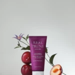 Purple Real Prune color-protecting treatment tube surrounded by fresh prunes and green leaves against a pale background.