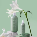 Green-tinted shampoo and scalp serum bottles with white lilies against a mint green background, beauty product shoot for Real Tamanu