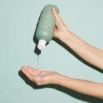 Hand holding a pastel green squeeze bottle and squeezing out lotion onto the other palm against a mint background.