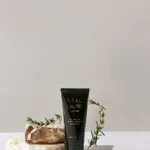 Tube of Real Grow anti-hair loss shampoo standing upright on stacked rocks, with sprigs of rosemary as a natural prop.