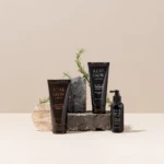 Black tubes of Real Grow anti-hair loss shampoo and treatment standing on a rock with rosemary sprigs against a beige backdrop.