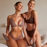 Two women in matching brown lingerie smile while holding gray bottle skincare products labeled BALIBODY on a bed.