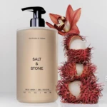 Beige Salt & Stone body wash bottle with pump, label reads 'Salt & Stone' and 'Saffron & Cedar', beside a red rambutan fruit with white interior and a pink flower.