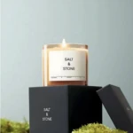 Candle labeled 'Salt & Stone' atop a black box with its lid slightly open, moss in foreground.