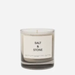 Scented candle in a clear glass jar with a beige label reading 'SALT & STONE' and a single wick visible on top.