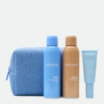 Three sunscreen bottles in blue and tan colors stand upright in front of a textured blue towel on a light background.