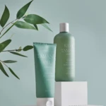 Green Tamanu oil bottles and tube displayed on white blocks with a leafy branch against a teal background (soothing scalp care product).
