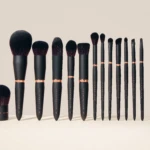 Row of black-handled makeup brushes with copper ferrules on a pale beige background, arranged from largest to smallest to the right.