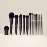 Row of black makeup brushes with copper accents arranged from largest to smallest on a beige background, showcasing a full brush set.