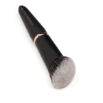 Close-up of a black makeup brush with a rose-gold ferrule and soft gray-bristled tip on a white background, angled diagonally.