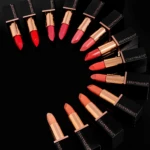 Row of coral and pink lipsticks with gold caps arranged diagonally on a black surface, with Youngblood packaging nearby.