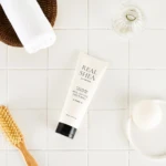 Flat lay of skincare items on white tiles: a diagonal tube labeled Real Shea among a wooden brush and a white towel in a basket area and a glass bottle nearby.