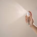 Hand holding a spray bottle releasing a fine mist toward a light beige wall.