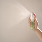 Hand holding a spray bottle spraying mist against a beige wall.