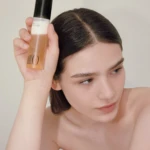 Woman with dark hair holds a two-tone cosmetic bottle near her temple, showing the product against her hairline.