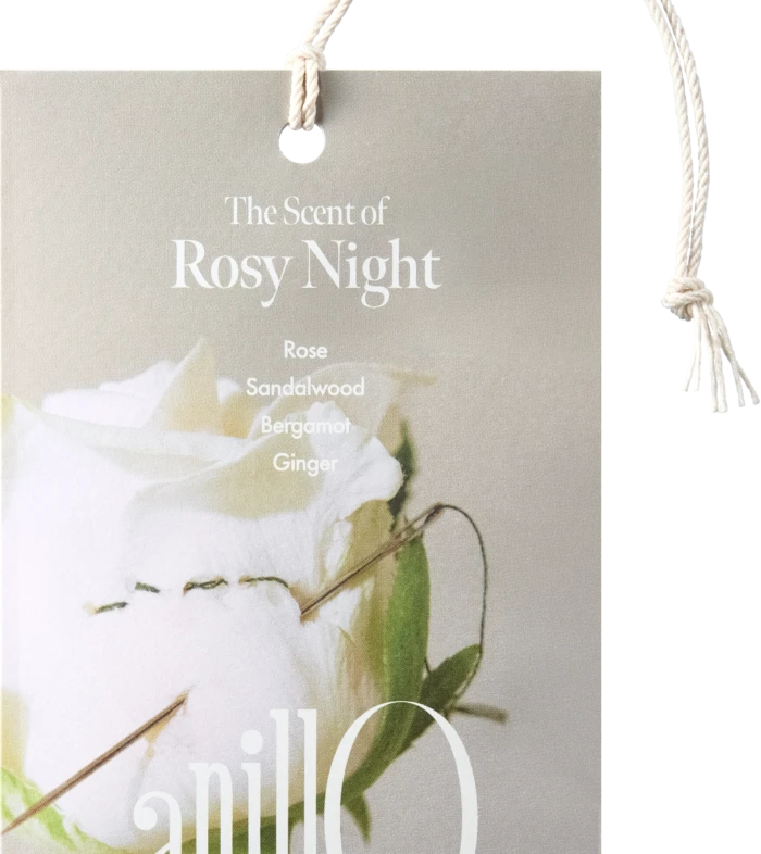 Beige gift tag with a white rope handle reading 'The Scent of Rosy Night' and listing notes: Rose, Sandalwood, Bergamot, Ginger, with a floral image in the background